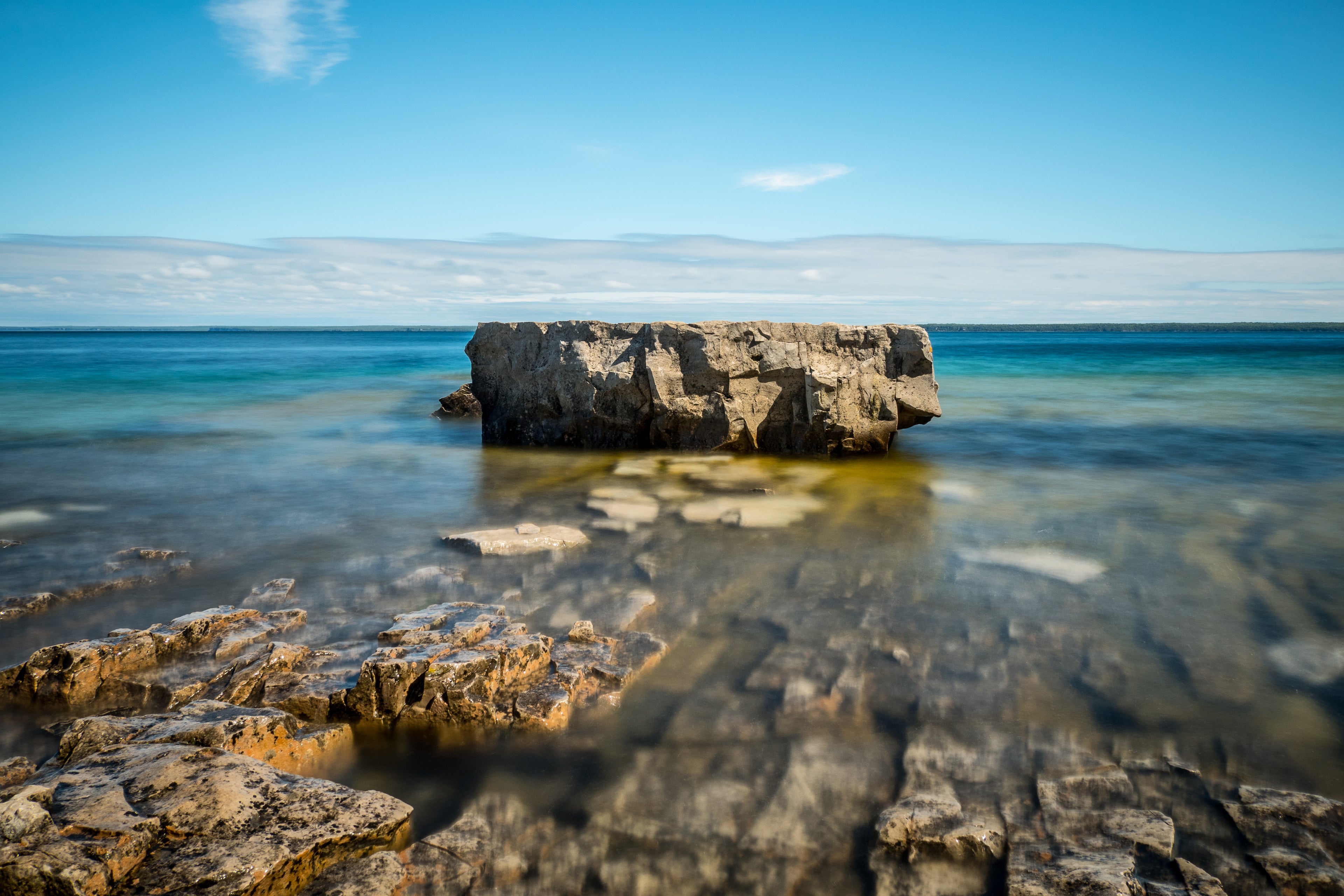 Image of a large, square rock in shallow, blue-green water. You can see the flat rocks of the shoreline in the foreground.