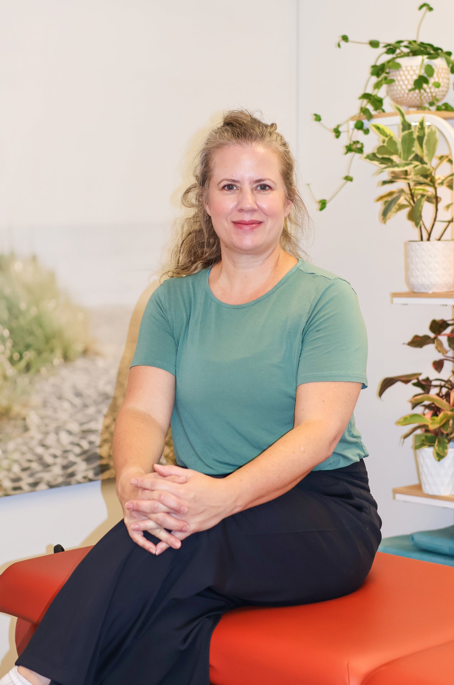 Picture of a woman, sitting on a massage table with one leg crossed over the other. Plants are visible in the background. 